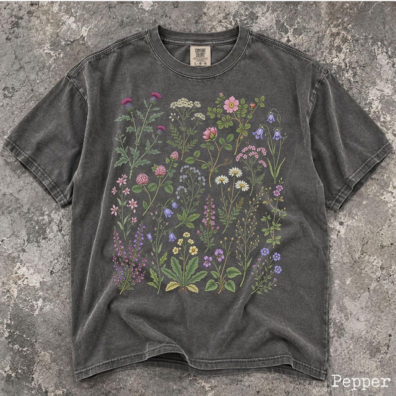 May include: A charcoal gray short-sleeve t-shirt featuring a detailed botanical print of various wildflowers in shades of pink, purple, yellow, and white. The shirt has a crew neckline and a Comfort Colors brand label printed on the inner collar.