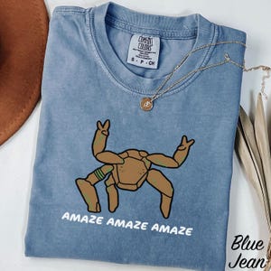 May include: A blue t-shirt with a graphic of a brown, multi-legged creature with raised arms. The text "AMAZE AMAZE AMAZE" is printed in white below the graphic. The shirt features a Comfort Colors label at the neck and a gold-toned necklace.