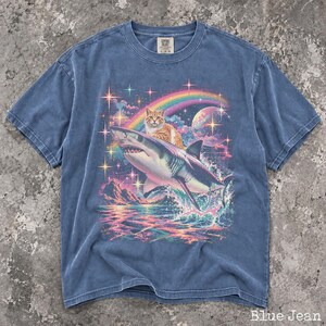 May include: A blue jean colored t-shirt featuring a graphic print of an orange tabby cat riding a shark. The background includes a rainbow, a moon, and colorful stars over a sea with mountains. The shirt has a crew neck and short sleeves.