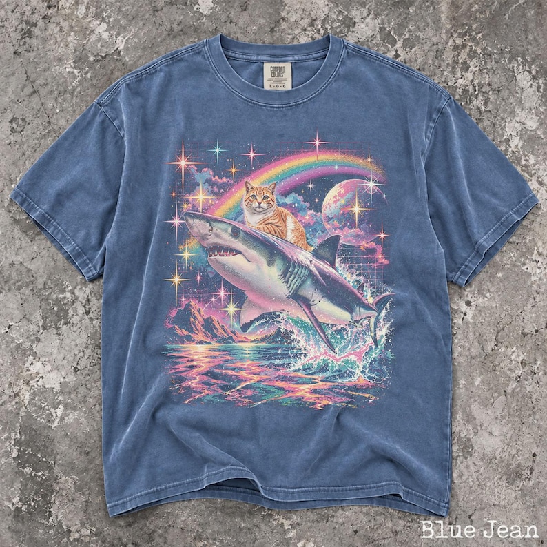 May include: A blue jean colored t-shirt featuring a graphic print of an orange tabby cat riding a shark. The background includes a rainbow, a moon, and colorful stars over a sea with mountains. The shirt has a crew neck and short sleeves.