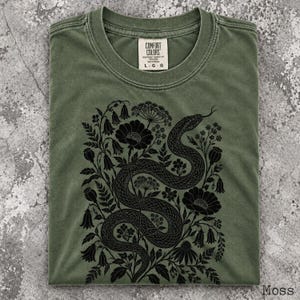 May include: An olive green crew neck t-shirt featuring a black graphic print of a coiled snake surrounded by various flowers and foliage. The interior neck label displays the text Comfort Colors and the size L.