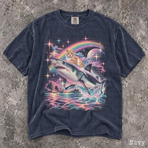 May include: A navy blue short-sleeve t-shirt featuring a graphic print of an orange tabby cat riding a shark over a colorful ocean scene. The design includes a rainbow, a moon, and sparkling stars against a dark sky with a grid pattern background.