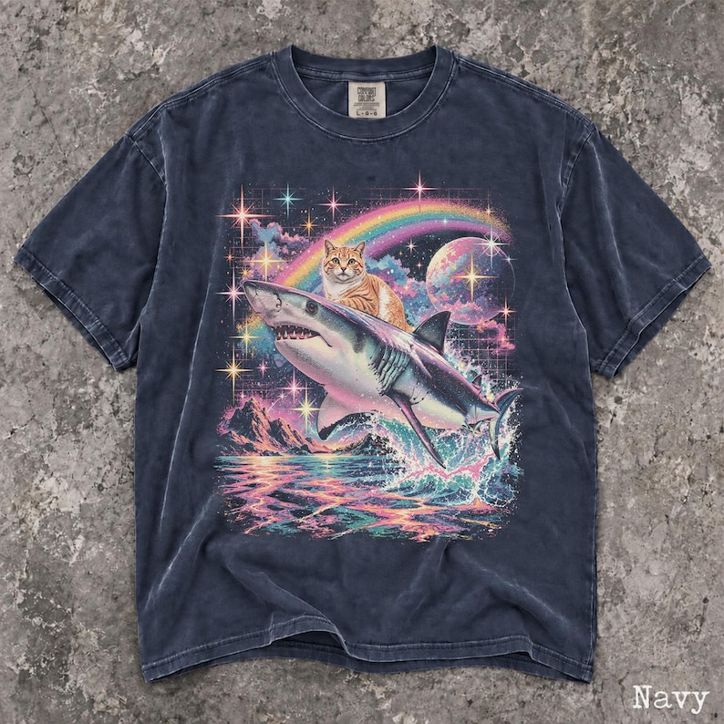 May include: A navy blue short-sleeve t-shirt featuring a graphic print of an orange tabby cat riding a shark over a colorful ocean scene. The design includes a rainbow, a moon, and sparkling stars against a dark sky with a grid pattern background.