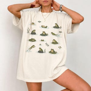 May include: An off-white short-sleeve t-shirt featuring a repeating graphic print of various green frogs in different poses, including sitting on lily pads, swimming, and resting on rocks, arranged in a grid-like pattern across the front.