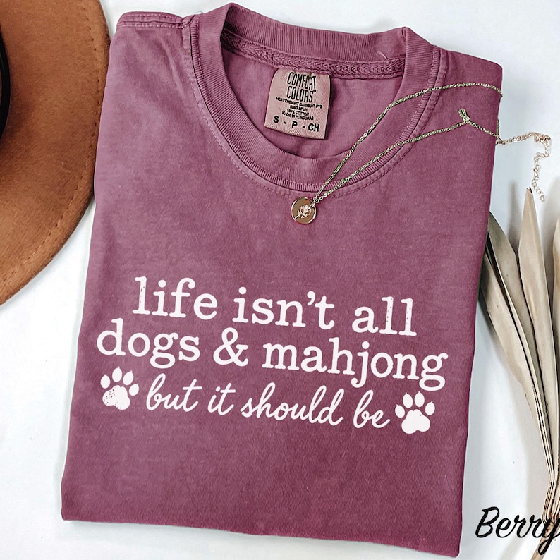 May include: A berry-colored short-sleeve t-shirt with a crew neckline and a Comfort Colors label. The shirt features white text that reads, life isn't all dogs & mahjong but it should be, accompanied by two white paw print graphics on the front.