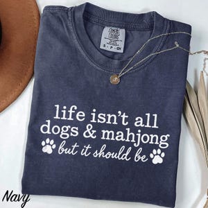 May include: A navy blue short-sleeve t-shirt with white text that reads "life isn't all dogs & mahjong but it should be" along with two white paw print graphics. The shirt features a crew neckline and a Comfort Colors brand label at the neck.