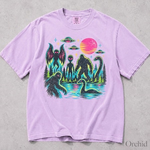 May include: A light purple short-sleeve t-shirt featuring a graphic print of a forest scene with a moth-like creature, an alien, a sasquatch, and a sea monster near a lake, under a pink sun and flying saucers, with the text Comfort Colors at the neck.
