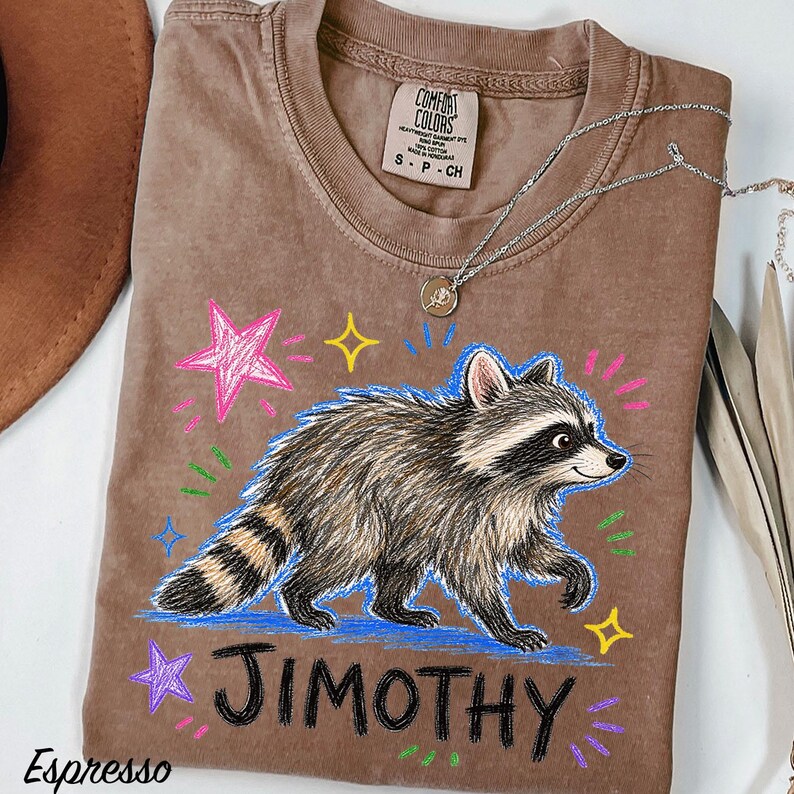 May include: A brown t-shirt featuring a graphic of a raccoon walking, surrounded by colorful hand-drawn style stars and dashes. The name JIMOTHY is printed in black below the raccoon. The shirt has a Comfort Colors label showing size S.