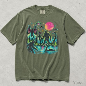 May include: An olive green short-sleeve t-shirt featuring a graphic print of cryptids including Bigfoot, a moth-like creature, and a sea serpent, alongside an alien, flying saucers, a pink sun, and a forest landscape against a dark background.