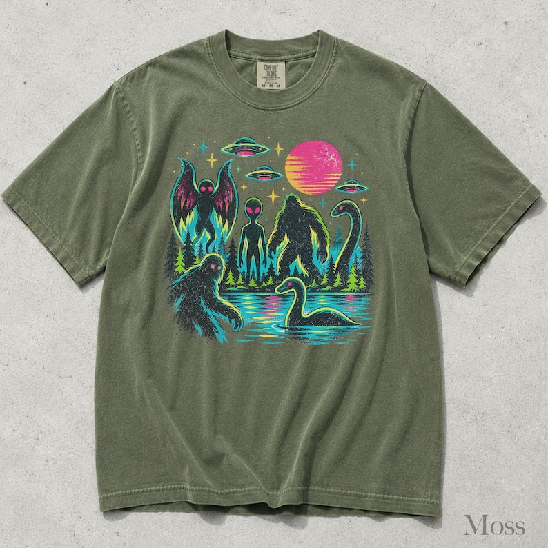 May include: An olive green short-sleeve t-shirt featuring a graphic print of cryptids including Bigfoot, a moth-like creature, and a sea serpent, alongside an alien, flying saucers, a pink sun, and a forest landscape against a dark background.