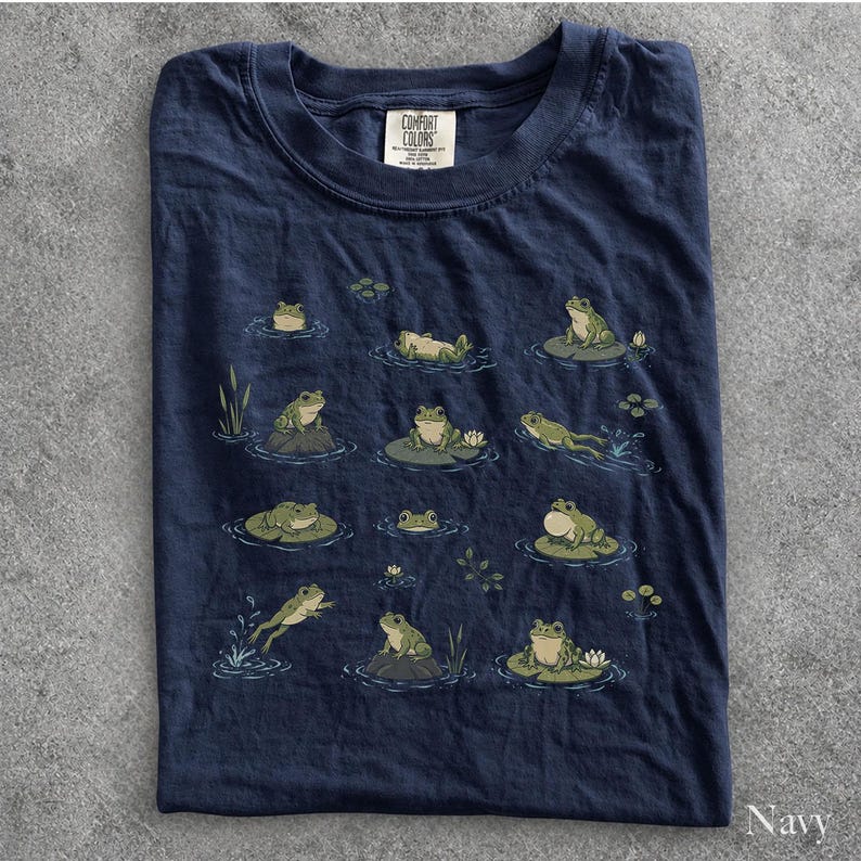 May include: A navy blue t-shirt featuring a graphic print of various green frogs in different poses, including sitting on lily pads, swimming, and resting on rocks. The shirt has a crew neckline and a Comfort Colors brand label at the inner collar.