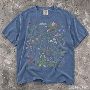 May include: A blue jean colored short-sleeve t-shirt featuring a detailed botanical print of various wildflowers in shades of pink, purple, white, and yellow. The shirt has a crew neckline and a Comfort Colors brand label at the inner collar.