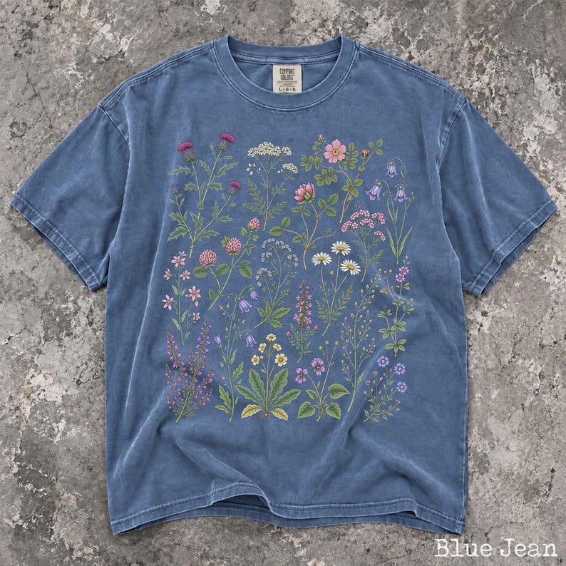 May include: A blue jean colored short-sleeve t-shirt featuring a detailed botanical print of various wildflowers in shades of pink, purple, white, and yellow. The shirt has a crew neckline and a Comfort Colors brand label at the inner collar.