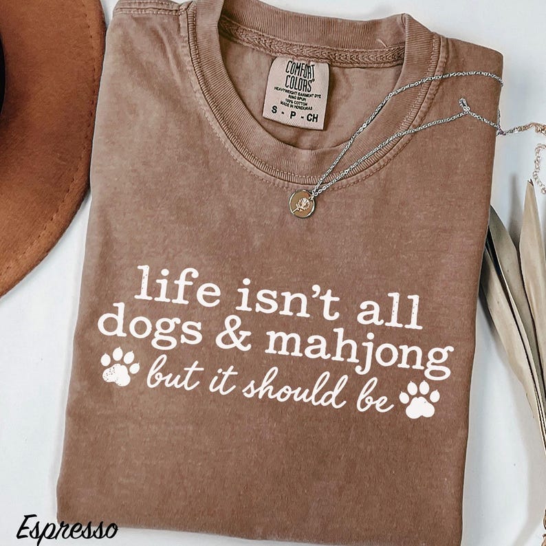May include: A brown short-sleeved t-shirt with white text that reads: life isn't all dogs & mahjong but it should be. The shirt features two white paw print graphics and a Comfort Colors brand label at the inner neckline. A silver necklace is visible.