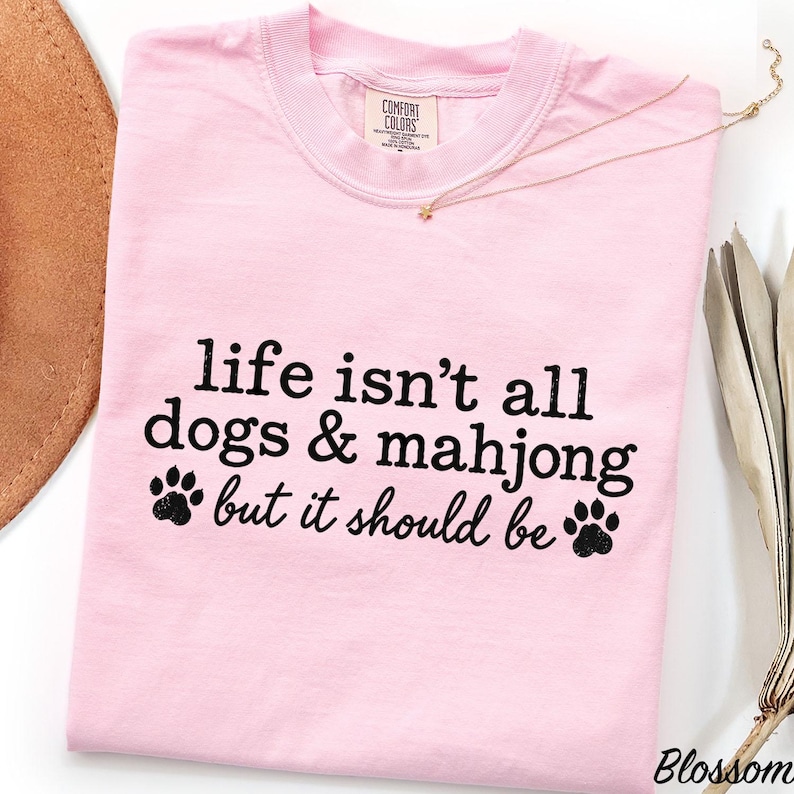 May include: A light pink short-sleeved t-shirt with a crew neckline. The front features black text that reads, "life isn't all dogs & mahjong but it should be," accompanied by two black paw print graphics, one on each side of the bottom line of text.