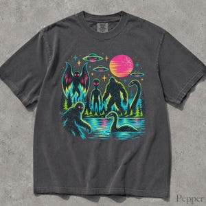 May include: A charcoal gray short-sleeve t-shirt featuring a vibrant graphic print of cryptids including Bigfoot, a moth-like creature, an alien, and a lake monster. The design includes neon-colored UFOs, a pink sun, stars, and a forest landscape.