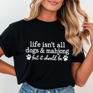May include: A black short-sleeved t-shirt featuring white text that reads, life isn't all dogs & mahjong but it should be, accompanied by two white paw print graphics positioned on either side of the bottom line of text.