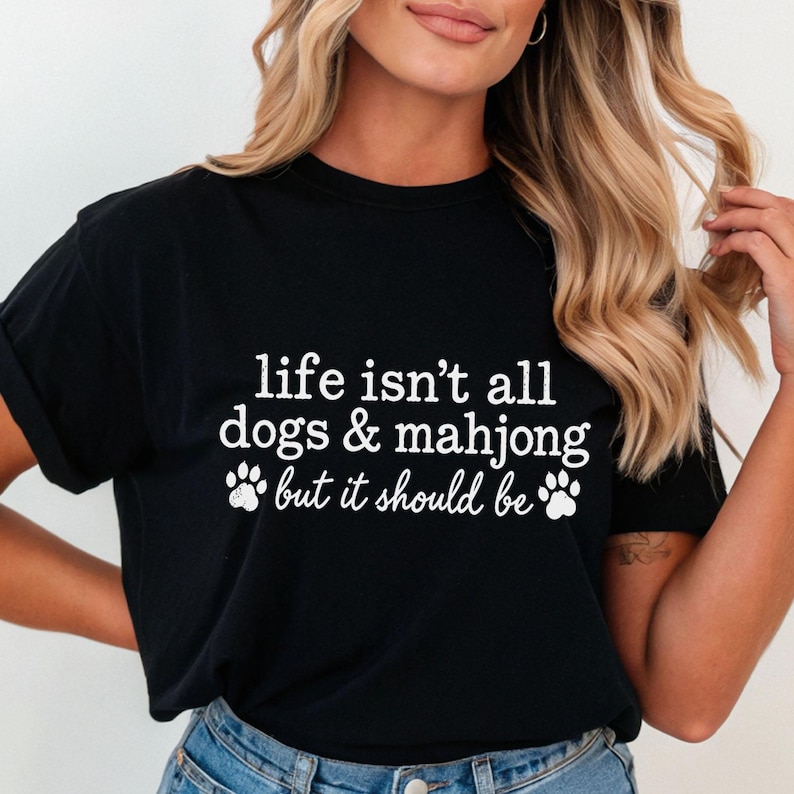 May include: A black short-sleeved t-shirt featuring white text that reads, life isn't all dogs & mahjong but it should be, accompanied by two white paw print graphics positioned on either side of the bottom line of text.
