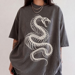 Puede incluir: Una camiseta de manga corta color gris carbón con un gran estampado gráfico en blanco de un esqueleto de serpiente enroscada. El diseño muestra a la serpiente con la boca abierta, colmillos afilados y una estructura espinal segmentada.