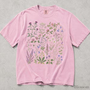 May include: A short-sleeved, crew-neck t-shirt in a faded pink color. The front features a detailed, multi-colored botanical illustration of various wildflowers, including thistles, daisies, and bell-shaped flowers, arranged in a dense, vertical cluster.