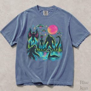 May include: A blue short-sleeve t-shirt featuring a graphic print of cryptids including Bigfoot, Mothman, and the Loch Ness Monster alongside an alien, flying saucers, and a pink sun over a forest and water landscape with a retro aesthetic.