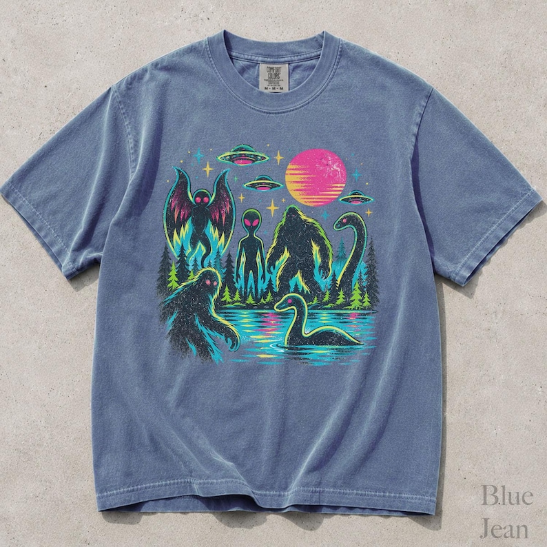 May include: A blue short-sleeve t-shirt featuring a graphic print of cryptids including Bigfoot, Mothman, and the Loch Ness Monster alongside an alien, flying saucers, and a pink sun over a forest and water landscape with a retro aesthetic.