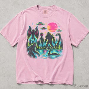 May include: A light pink short-sleeve t-shirt featuring a graphic print of a forest scene with a moth-like creature, an alien, a sasquatch, and a sea monster near water. Above the trees are three flying saucers and a bright pink sun.