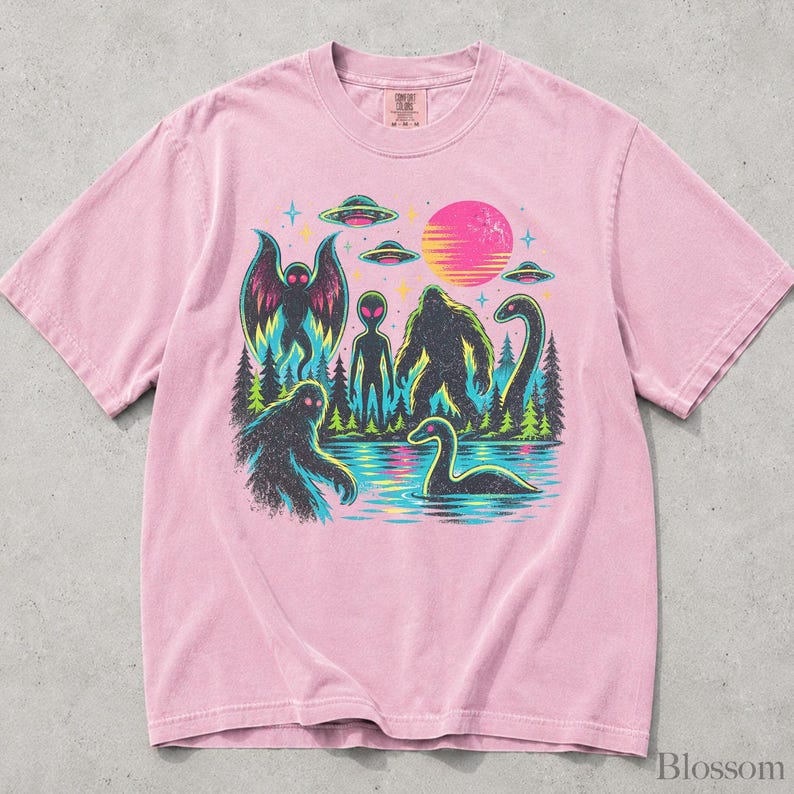 May include: A light pink short-sleeve t-shirt featuring a graphic print of a forest scene with a moth-like creature, an alien, a sasquatch, and a sea monster near water. Above the trees are three flying saucers and a bright pink sun.