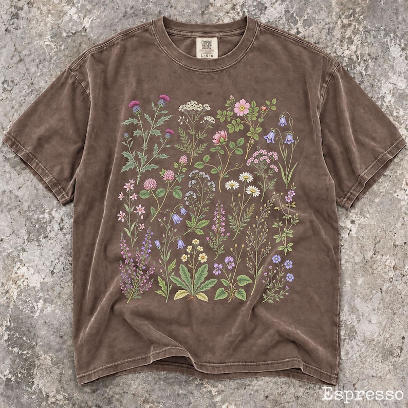 May include: A brown short-sleeve t-shirt featuring a colorful botanical print of various wildflowers and plants across the front. The shirt has a crew neckline and a label at the inner neck area. The fabric has a washed, vintage-style appearance.