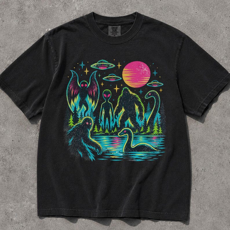May include: A black short-sleeve t-shirt featuring a vibrant graphic print of cryptids and extraterrestrial elements. The design includes a moth-like creature, an alien, a sasquatch, and a sea monster near a lake, set against a forest and neon sun.