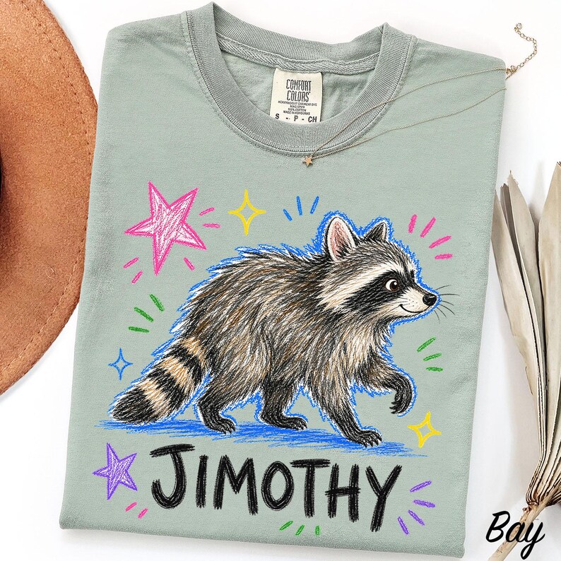 May include: A sage green t-shirt featuring a detailed illustration of a raccoon walking. The design includes colorful hand-drawn style stars and dashes in pink, yellow, and blue. The text JIMOTHY is printed in a bold, black, hand-lettered font below.