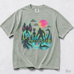 May include: A sage green short-sleeve t-shirt featuring a graphic print of cryptids including Bigfoot, a moth-like creature, and a sea serpent, alongside an alien, flying saucers, a forest, and a pink sun, with a Comfort Colors brand label.