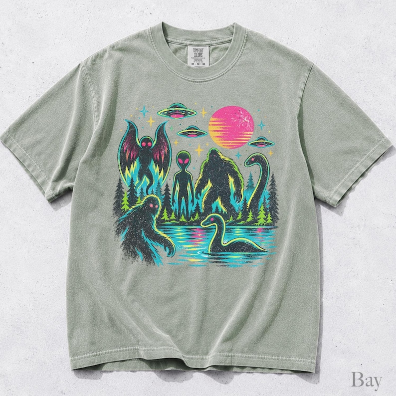 May include: A sage green short-sleeve t-shirt featuring a graphic print of cryptids including Bigfoot, a moth-like creature, and a sea serpent, alongside an alien, flying saucers, a forest, and a pink sun, with a Comfort Colors brand label.