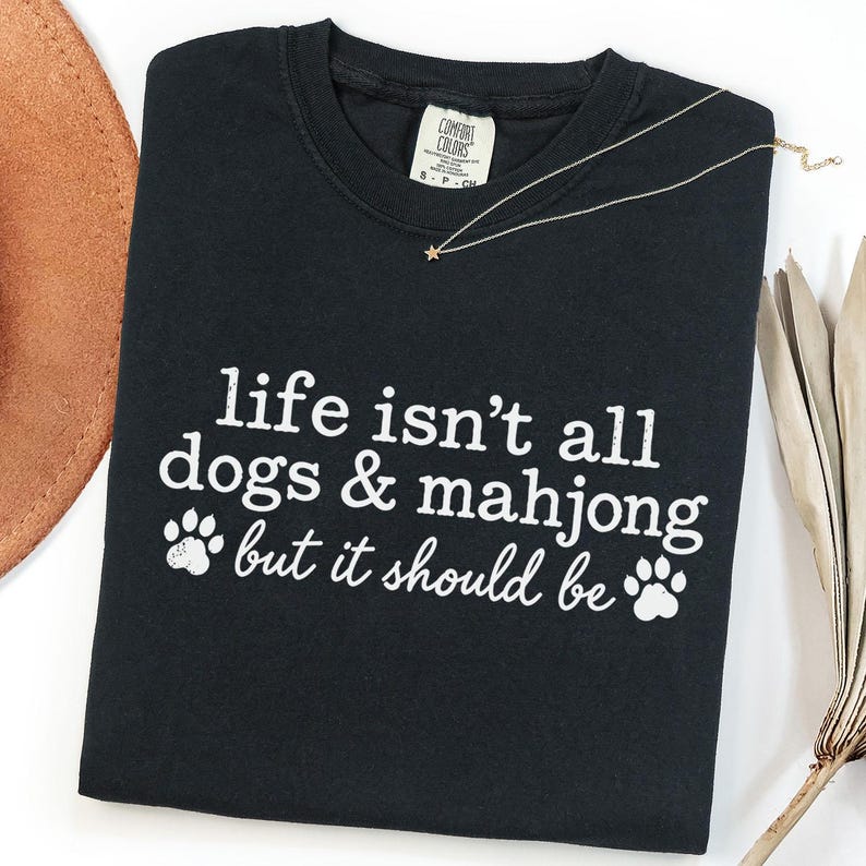 May include: A black crewneck t-shirt featuring white screen-printed text that reads, "life isn't all dogs & mahjong but it should be," accompanied by two white paw print graphics. The shirt has a Comfort Colors brand label at the inner neckline.