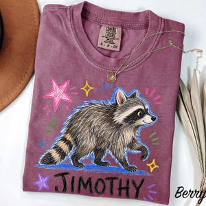 May include: A berry-colored t-shirt featuring a graphic of a raccoon with a striped tail. The design includes colorful hand-drawn style stars and the name JIMOTHY printed in black. The shirt has a Comfort Colors label at the inner neckline.