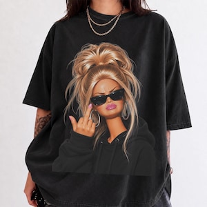 Puede incluir: Una camiseta negra de manga corta con un estampado gráfico de una persona rubia con un moño alto, gafas de sol negras y una sudadera con capucha negra. El gráfico muestra a la persona haciendo un gesto con el dedo corazón levantado.