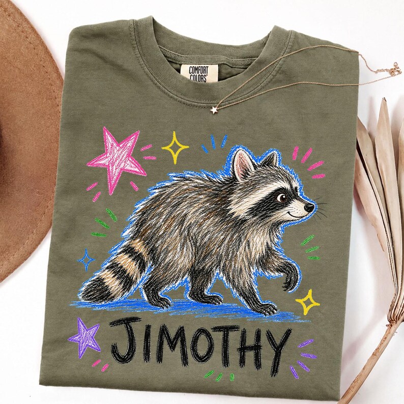 May include: An olive green t-shirt featuring a graphic of a raccoon with a striped tail. The design includes colorful hand-drawn style stars and dashes in pink, yellow, and blue. The name JIMOTHY is printed in black below the raccoon illustration.