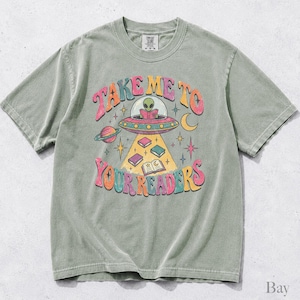 May include: A sage green short-sleeve t-shirt featuring a graphic of an alien in a flying saucer reading a book. The text reads "TAKE ME TO YOUR READERS" in colorful, wavy letters. The design includes planets, stars, and books floating in a beam.