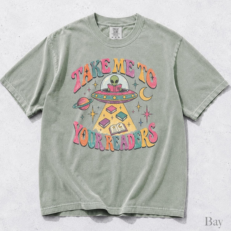 May include: A sage green short-sleeve t-shirt featuring a graphic of an alien in a flying saucer reading a book. The text reads "TAKE ME TO YOUR READERS" in colorful, wavy letters. The design includes planets, stars, and books floating in a beam.