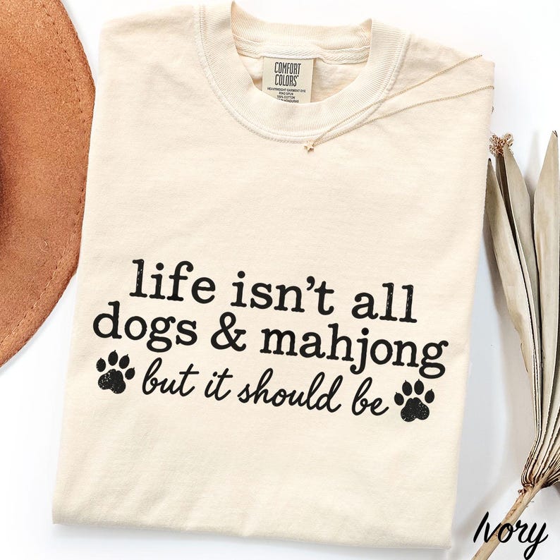 May include: An ivory-colored crew neck t-shirt featuring black text that reads, "life isn't all dogs & mahjong but it should be," accompanied by two black paw print graphics. The shirt has a Comfort Colors brand label at the inner neckline.