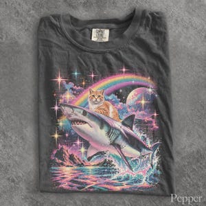 May include: A charcoal gray t-shirt featuring a graphic print of an orange tabby cat riding a shark. The background displays a colorful rainbow, a large planet, sparkling stars, and a mountain landscape over water with a grid pattern.