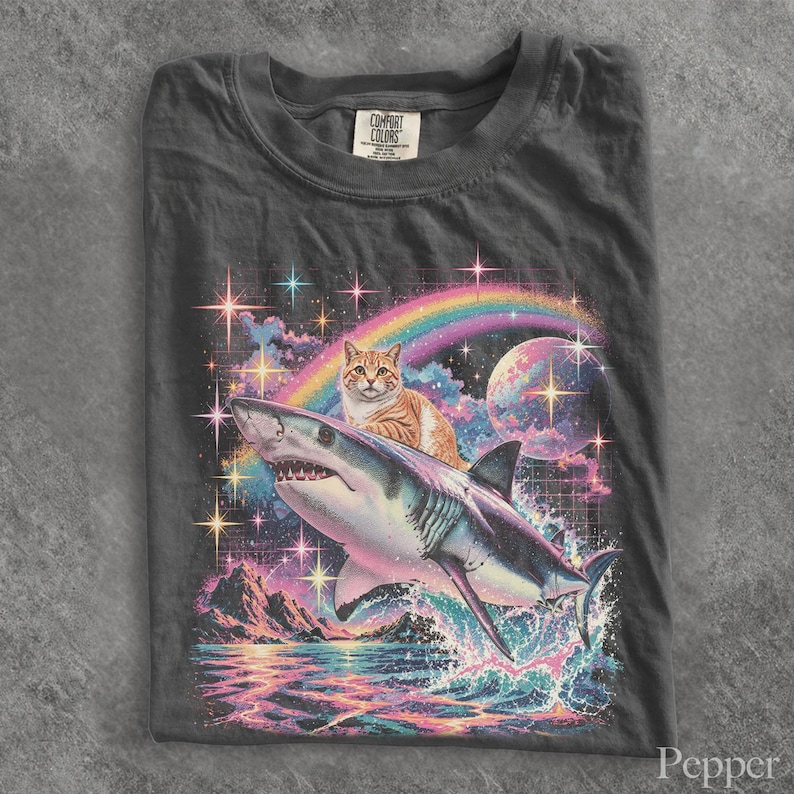 May include: A charcoal gray t-shirt featuring a graphic print of an orange tabby cat riding a shark. The background displays a colorful rainbow, a large planet, sparkling stars, and a mountain landscape over water with a grid pattern.