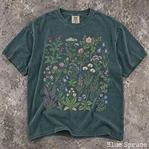 May include: A dark teal, pigment-dyed short-sleeve t-shirt featuring a detailed botanical print of various wildflowers in shades of pink, purple, yellow, and white. The shirt has a crew neckline and a visible brand label at the inner collar.