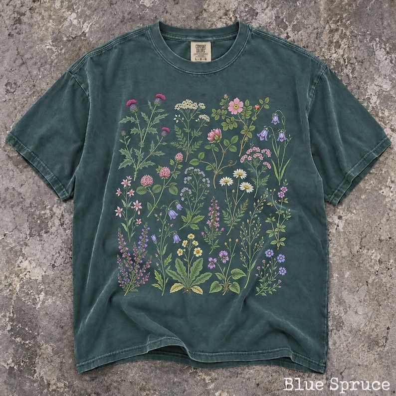 May include: A dark teal, pigment-dyed short-sleeve t-shirt featuring a detailed botanical print of various wildflowers in shades of pink, purple, yellow, and white. The shirt has a crew neckline and a visible brand label at the inner collar.