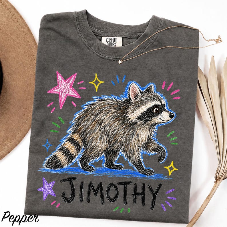 May include: A charcoal gray t-shirt featuring a graphic of a raccoon with a striped tail, surrounded by colorful hand-drawn style stars and dashes. The name JIMOTHY is printed in a bold, black, hand-lettered font below the raccoon illustration.