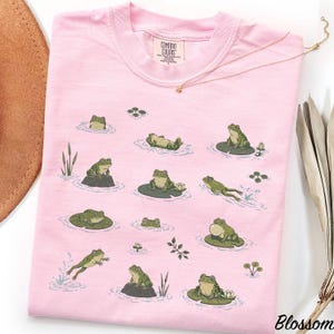 May include: A light pink t-shirt featuring a repeating graphic print of green frogs in various poses, including sitting on lily pads, swimming, and resting on rocks. The shirt has a crew neckline and a Comfort Colors brand label at the inner neck.