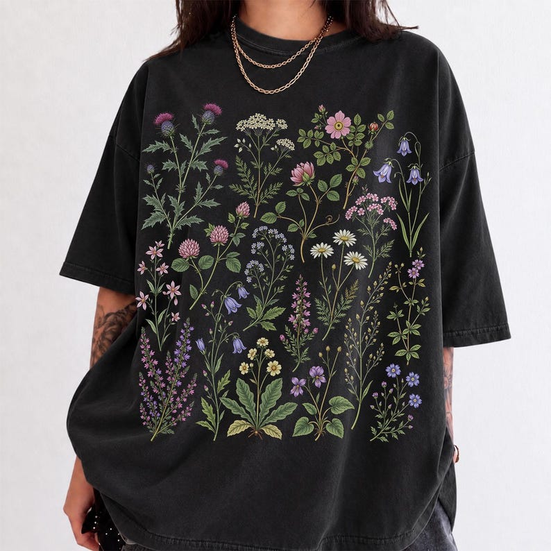 May include: A black short-sleeve t-shirt featuring a detailed botanical print of various wildflowers and plants in shades of pink, purple, blue, white, and green. The graphic covers the center of the shirt, displaying diverse floral species.