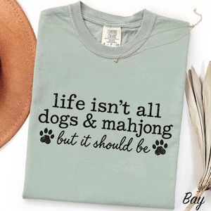 May include: A sage green crew neck t-shirt featuring the black text "life isn't all dogs & mahjong but it should be" with two paw print graphics. The shirt has a Comfort Colors brand label at the inner neckline and a short-sleeved design.