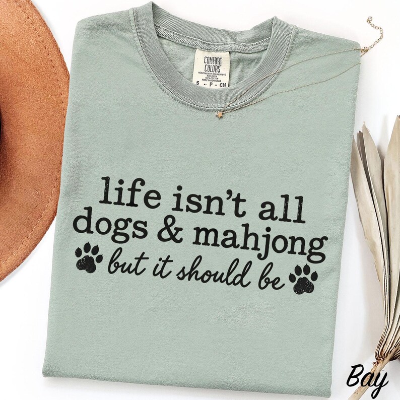 May include: A sage green crew neck t-shirt featuring the black text "life isn't all dogs & mahjong but it should be" with two paw print graphics. The shirt has a Comfort Colors brand label at the inner neckline and a short-sleeved design.