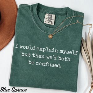May include: A dark green short-sleeve t-shirt with a crew neckline and a vintage-wash finish. The shirt features the white printed text: i would explain myself but then we'd both be confused. A gold-toned necklace with a small circular pendant rests on the shirt.